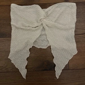 Vit bandeau tubtopp med knutdetalj - Säljer en vit bandeau tubtopp med snygg knutdetalj framtill och spetsiga kanter nedtill. Toppen har ett vågigt, strukturerat tyg som ger en cool look. Perfekt för dig som vill sticka ut med en unik och trendig topp i sommar.