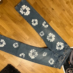 Levi's 501 x Denim Tears jeans med blommor - Unika Levi's 501 jeans i samarbete med Denim Tears, dekorerade med vita blommiga tryck över hela byxorna. Klassisk blå denim, rak passform och ikoniska detaljer som röd Levi's-tag och patch bak. Perfekta för dig som vill sticka ut med en kreativ och trendig look.