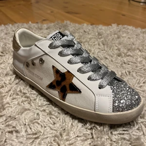Golden Goose sneakers med glitter och leopard - Snygga vita Golden Goose sneakers med silvriga glittriga snören och tå, leopardmönstrad stjärna på sidan och gulddetalj vid hälen. Skorna har rund tå, platt sula och coola metalldetaljer. Perfekta för dig som vill sticka ut med din stil.