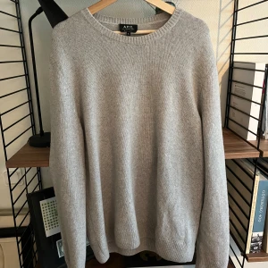 Ljusgrå stickad tröja från A.P.C. - Säljer en stilren ljusgrå stickad tröja från A.P.C. i storlek XL. Tröjan är tillverkad i en lyxig mix av 50% ull och 50% alpacka, har rund halsringning och långa ärmar. Perfekt för dig som gillar minimalistisk och clean look.