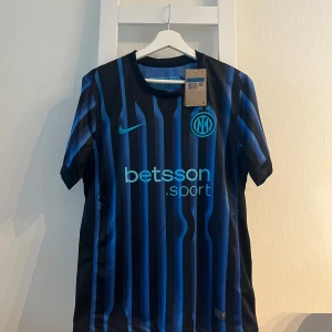 Inter Milan randig fotbollströja Nike - Snygg Inter Milan fotbollströja från Nike med svarta och blå vertikala ränder, klubbmärke och Nike-logga på bröstet samt sponsortryck. Tillverkad i lätt och ventilerande polyester, perfekt för match eller träning.