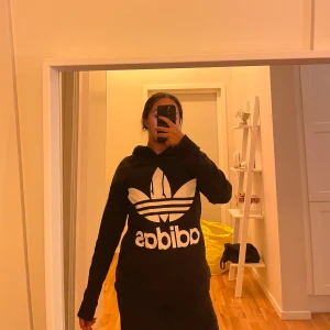 Svart Adidas hoodie med stor logga - Svart hoodie från Adidas med stor vit trefoil-logga och text på bröstet. Klassisk huva med snörning och långa ärmar. Perfekt för en avslappnad och sportig stil.