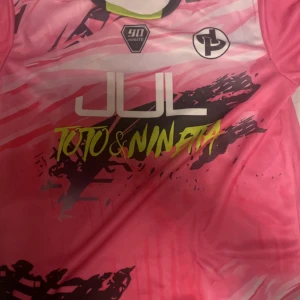 Rosa fotbollströja JUL x Toto & Ninetta - Snygg fotbollströja i rosa med grafiskt mönster och tryck från JUL x Toto & Ninetta. Tröjan har korta ärmar, svart och vit logga på bröstet och coola detaljer i neongult och svart. Perfekt för dig som vill sticka ut på planen.