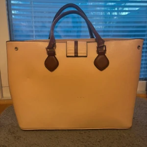 Beige handväska med bruna detaljer - Stilren beige handväska med bruna handtag och detaljer. Väskan har en rymlig insida med flera fack och är tillverkad i syntetmaterial med struktur. Perfekt för dig som vill ha en klassisk och enkel väska med plats för allt du behöver.