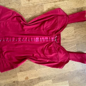 Vintage Röd korsett-topp från Guess Jeans - Snygg vintage röd korsett-topp från Guess Jeans med breda axelband och dekorativa krokar framtill. Toppen har Guess-logga i strass på sidan och tryckt emblem på insidan. Figurnära passform och lätt glansigt bomullsmaterial. Perfekt för dig som vill sticka ut.