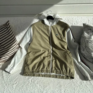 Lacoste vindjacka - Snygg vindjacka från Lacoste Sport i beige och vitt med huva och hel dragkedja. Jackan har vita ärmar med diskreta mönsterdetaljer längs sidorna och klassisk Lacoste-logga på bröstet. Regular fit och elastisk nederkant för en sportig vibe.