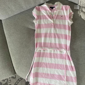 Randig kort klänning Tommy Hilfiger - Superfin kort barn klänning från Tommy Hilfiger i rosa och vitt med breda ränder. Klassisk pikékrage, knappar framtill och knytskärp i midjan. Mjuk bomullskvalitet och kortärmad modell – perfekt för en fräsch och sportig look. Ålder 8-9 år.