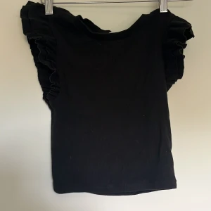 Svart volangtopp från Zara - Svart topp från Zara med volangdetaljer längs axlarna. Modellen är ärmlös och har en rund halsringning. Toppens material är ribbad bomull som ger en skön och stretchig känsla. Perfekt för dig som gillar en stilren look med en twist.