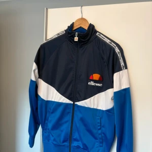 Ellesse blå och vit track jacket S - Unik track jacket från Ellesse i marinblått, blått och vitt med logga på bröstet och Ellesse-band längs axlarna. Jackan har dragkedja framtill, ståkrage och två fickor. Perfekt för en sportig och retro vibe.