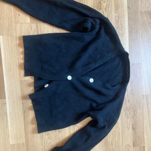 Svart stickad kofta med knappar - Mysig svart stickad kofta med v-ringning och tre vita knappar framtill. Koftan har långärmad design och ribbade muddar, perfekt för lager-på-lager-stil. Enkel och stilren modell som passar till det mesta.