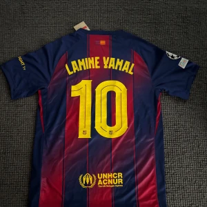 FC Barcelona Lamine Yamal tröja Nike L - Säljer en officiell FC Barcelona matchtröja från Nike med Lamine Yamal och nummer 10 på ryggen. Tröjan är blå och röd med gula detaljer, randigt mönster, klubbmärke, Champions League-märke och sponsorer som Spotify och UNHCR. Tillverkad i Dri-FIT-material.