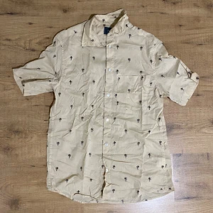 Beige kortärmad skjorta H&M palmtryck - Snygg beige skjorta från H&M med svart palmtryck över hela. Regular fit med korta ärmar, klassisk krage och bröstficka. Perfekt för chill sommardagar eller när du vill ha en avslappnad vibe. Gjord i mjuk bomull som andas.