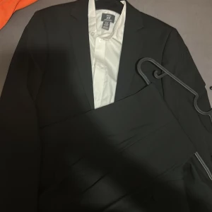 Svart kostym från H&M med vit skjorta - Snygg svart kostym från H&M med klassisk kavaj och matchande kostymbyxor. Kavajen har två knappar och smal krage, och till kostymen ingår en vit skjorta. Materialet är polyester och passformen är normal. Perfekt för dig som vill ha en stilren och enkel look.