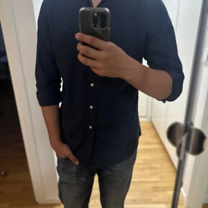 Mörkblå skjorta från Polo Ralph Lauren - Snygg mörkblå skjorta från Polo Ralph Lauren med klassisk button-down krage och broderad blå logga på bröstet. Skjortan har långa ärmar och vita knappar framtill. Tillverkad i mjuk bomull för en skön känsla.