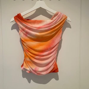 Färgstark omlott topp i mesh - Säljer en snygg ärmlös topp i mesh med draperad effekt och omlott-detalj. Toppen har en mix av orange, rosa och vit färg i ett mjukt, nästan akvarell-liknande mönster. 