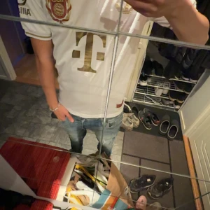 Vit Bayern München Adidas fotbollströja - Snygg vit Bayern München fotbollströja från Adidas med gulddetaljer och klubbens 125-årsjubileumsmärke på bröstet. Tröjan har korta ärmar, klassisk rund hals och är tillverkad i lätt funktionsmaterial (AEROREADY) som andas. Perfekt för dig som älskar fotboll och stil.