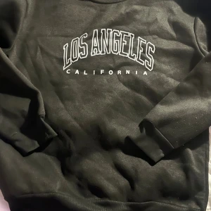 Svart sweatshirt Los Angeles M - Svart sweatshirt med trycket 'Los Angeles California' i vitt framtill. Tröjan har rund hals, långa ärmar och är tillverkad i mjuk polyester. Perfekt för en chill och avslappnad stil.