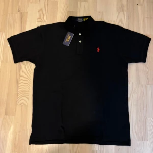 Ralph Lauren Polo tröja svart - Svart pikétröja från Polo Ralph Lauren med klassisk krage, tre knappar och röd broderad logga på bröstet. Tröjan är i bomull och har en slim passform som sitter snyggt. Perfekt för dig som gillar stilren och tidlös design.