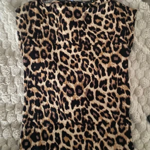 Leopardmönstrad ärmlös topp - Snygg ärmlös topp med leopardmönster i svart och bruna toner på beige botten. Modellen har rund halsringning och rak passform, perfekt för dig som vill sticka ut med ett djurmönstrat plagg. Materialet känns mjukt och följsamt mot huden.