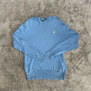 Blå stickad tröja Polo Ralph Lauren S - Blå stickad tröja från Polo Ralph Lauren i slim fit med klassisk gul broderad logga på bröstet. Långärmad modell med rund hals och ribbade muddar. Perfekt för lager-på-lager och snygg till jeans eller chinos.