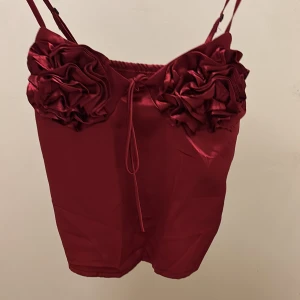 Vinröd satin linnetopp med rosetter - Supersnygg vinröd linnetopp i satin med smala axelband och stora dekorativa rosetter framtill. Toppen har en glansig finish och justerbara axelband för perfekt passform. Perfekt för dig som vill sticka ut med en unik detalj.