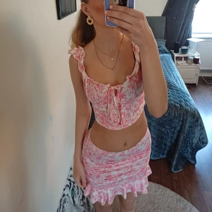 Blommig croppad blus med volang och en tjol  - Supersöt blus i vitt och rosa med blommigt mönster. Toppen har volangdetaljer vid axelbanden, knyt framtill och fyrkantig rygg.ä och en tjol, det är ett set. Materialet känns lätt och somrigt, perfekt för dig som gillar romantisk stil och vill sticka ut.  Storlek S. Detet är från shein 