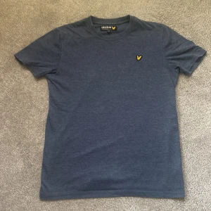 Blå t-shirt från Lyle & Scott Junior - Snygg blå t-shirt från Lyle & Scott Junior med klassisk rund hals och korta ärmar. T-shirten har den ikoniska gula örnloggan broderad på bröstet. Perfekt basic-plagg med skön passform och mjukt bomullsmaterial.stl 14/15
