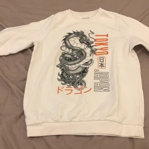 Vit sweatshirt med draktryck från Primark - Vit sweatshirt från Primark med stort svart draktryck och japanska tecken på framsidan. Texten 'Tokyo Japan' i orange och svart ger en cool streetstyle-vibe. Tröjan har rund hals och långa ärmar, perfekt för dig som gillar asiatiska influenser.