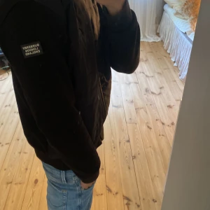 Svart Burberry cardigan  - Svart cardigan från Burberry med huva och dragkedja framtill. Jackan har ett broderat Burberry-emblem på bröstet och en patch på ärmen. Snygg, stilren design med ribbade muddar och fickor med dragkedja. Perfekt för dig som vill ha en clean look till hösten och även perfekt till våren.