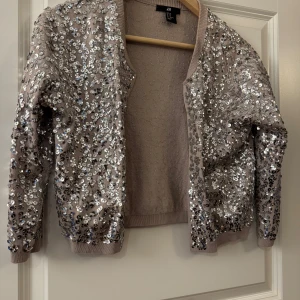 Glittrig kofta med paljetter från H&M - Säljer en  kofta från H&M som är täckt med silvriga paljetter. Koftan har rund halsringning, ribbade muddar och långa ärmar. Perfekt för dig som vill sticka ut och addera extra bling till din outfit!