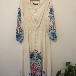 Lång blommig klänning med knappar - Långklänning i ljus beige med v-ringning och knappar framtill. Klänningen har långa ärmar med blommigt mönster i blått, rosa och grönt samt smock i midjan. Materialet känns lätt och flowy, perfekt för vår och sommar.