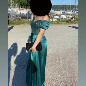 Grön offshoulder balklänning i satin - Elegant lång grön balklänning i glansigt satinmaterial med offshoulder-ärmar och figurnära passform. Livet är dekorerat med små pärlor och ger en lyxig känsla. Perfekt för bal eller speciella tillfällen.