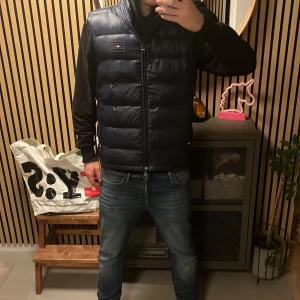 Mörkblå dunväst från Tommy Hilfiger - Snygg mörkblå dunväst från Tommy Hilfiger med quiltad design och dragkedja framtill. Västen har hög krage och diskret logga på bröstet. Perfekt för lager-på-lager och enkel att packa ner tack vare TH Packable Series. Materialet är polyester. Den är använd 2-3 gånger och är i väldigt bra skick. Ny kostar runt 2000kr men säljer den för 1200, hör gärna av er vid frågor och funderingar kan sänka priset vid snabbt köp.