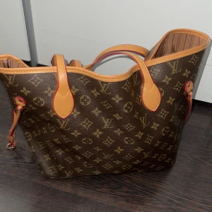Louis Vuitton Monogram axelväska - Säljer en klassisk Louis Vuitton Neverfull axelväska i brunt monogrammönster med beige och röda detaljer. Väskan har randigt innerfoder, dragkedjeficka och långa handtag. Tillverkad i slitstarkt skinn och canvas, perfekt för dig som gillar lyxig och tidlös design. Passar till allt. Simpel outfit men också för lite mer finklädd. Rymmer otroligt mycket. 💓säljer då den inte kommer till användning. Nypris 5000🫶🏼Skriv vid intresse! 