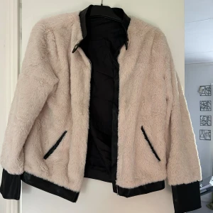 Beige teddyjacka med svarta detaljer - Säljer en beige teddyjacka med mjuk pälsimitation och svarta detaljer vid krage, muddar och fickor. Jackan har dragkedja framtill och är helfodrad. Perfekt för dig som vill ha en mysig och trendig look.