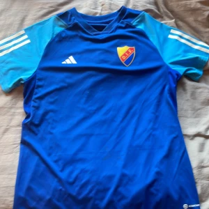 Blå Djurgården fotbollströja Adidas L - Djurgården fotbollströja från Adidas i storlek L. Tröjan är blå med ljusblå korta ärmar och vita Adidas-ränder på axlarna. DIF-emblemet på bröstet i gult, rött och blått. Tillverkad i lätt och ventilerande polyester, perfekt för träning eller match.