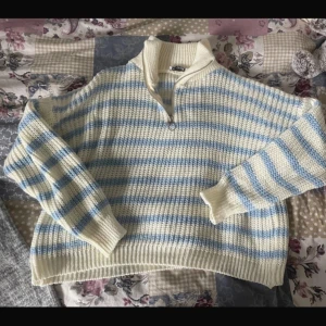 Randig stickad tröja Design by Si - Supermysig stickad tröja från Design by Si i vitt och ljusblått randigt mönster. Tröjan har hög krage med half zip-dragkedja och långa ärmar. Perfekt för kyliga dagar och ger en chill vibe till din outfit. ganska kort och kragen är i ett tjockare material 