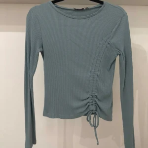 Blå/turkos långärmad topp med snörning - Säljer en ljusblå ribbad långärmad topp med snörning och rynkdetalj på sidan som kan spännas hur mycket eller lite man vill för olika stilar. Toppen har rund halsringning och är tillverkad i ett mjukt, stretchigt material som sitter snyggt på kroppen. Perfekt för dig som gillar trendiga detaljer och enkel stil.