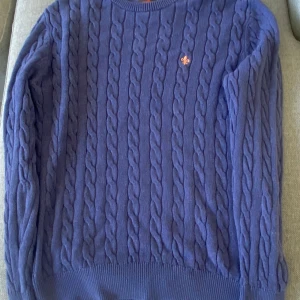 Blå kabelstickad tröja från Morris - Kabelstickad blå tröja från Morris i 100% pima bomull. Klassisk rund halsringning, ribbade muddar och diskret orange logga på bröstet. Perfekt för dig som gillar stilrena och tidlösa plagg med snygg struktur.