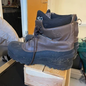 Bruna vinterkängor från BLMR - Robusta bruna kängor från BLMR med svart gummisula och snörning. Skorna är vattentäta och har en hög, vadderad krage i svart textil för extra komfort. Perfekta för kalla dagar och tuffare väder.