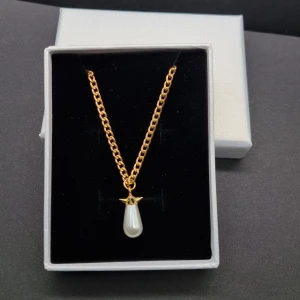 Guldfärgat halsband med pärlhänge - Stilrent halsband med guldfärgad kedja och ett droppformat vitt pärlhänge. Kedjan har klassisk länkdesign och hänget är dekorerat med en liten guldfärgad detalj upptill. Perfekt för dig som gillar en elegant och tidlös look.