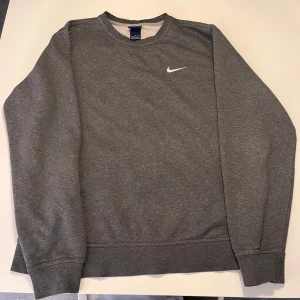 Grå Nike sweatshirt i fleece - Mysig grå sweatshirt från Nike med klassisk vit swoosh på bröstet. Tröjan är i mjuk fleece, har rund halsringning och långa ärmar. Perfekt för kalla dagar eller när du vill ha en enkel och stilren look.