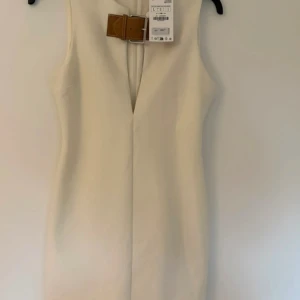 Beige ärmlös klänning med bälte - Stilren beige klänning med v-ringning och snyggt brunt bälte i midjan. Ärmlös modell med rak passform som ger en clean och modern look. Perfekt för dig som gillar minimalistisk stil.