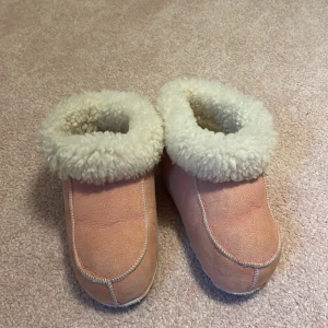 Rosa fårskinnstofflor med ullfoder - Mysiga tofflor i ljusrosa mocka med fluffigt vitt ullfoder. De har en platt sula i mocka och är perfekta för kalla dagar. Tofflorna har en enkel design med synliga sömmar och rund tå, och är supermjuka inuti för extra komfort. 