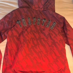 Röd Trapstar windbreaker  - Säljer en röd vindjacka från Trapstar med camo-mönster och stor logga på ryggen. Jackan har huva, dragkedja framtill och en snygg detalj med tryckt emblem på bröstet. Perfekt för dig som vill sticka ut med streetwear-vibe.