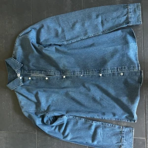 Blå jeansskjorta från Jack & Jones - Snygg blå jeansskjorta från Jack & Jones med klassisk krage och vita knappar framtill. Skjortan har lång ärm och är tillverkad i robust jeanstyg som ger en cool och avslappnad vibe. Perfekt att bära öppen över en t-shirt eller stängd för en mer clean look. Prislapp kvar.
