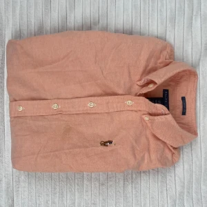 Ljus orange skjorta från Ralph Lauren - Snygg ljus orange skjorta från Ralph Lauren med klassisk krage och broderad polospelare på bröstet. Skjortan har långa ärmar, vita knappar och är gjord i mjuk bomull. Perfekt för dig som gillar stilrena och tidlösa plagg. Har en liten fläck under loggan (se bild 3) men inget som syns tydligt. Går förmodligen att ta bort i tvätten, men har inte testat. Passar XS-S