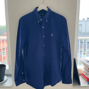 Blå skjorta från Ralph Lauren slim fit - Snygg blå skjorta från Ralph Lauren i slim fit med klassisk button-down krage och broderad logga på bröstet. Skjortan är tillverkad i 100% bomull med stretch och har vita knappar framtill och vid kragen. Perfekt för dig som gillar stilrena och tidlösa plagg.