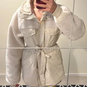 Beige teddyjacka med bälte - Säljer en beige teddyjacka med fluffiga ärmar och detaljer i teddyfleece. Jackan har knytbälte i midjan, stora bröstfickor och dragkedja framtill. Perfekt för dig som vill ha en mysig och trendig look under kyliga dagar.