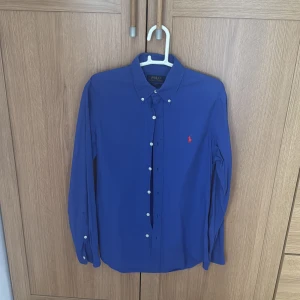 Blå skjorta från Polo Ralph Lauren - Snygg blå skjorta från Polo Ralph Lauren med klassisk button-down krage och vita knappar. Ikoniska röda loggan broderad på bröstet. Skjortan är långärmad och i mjuk bomull, perfekt för en clean och stilren look.